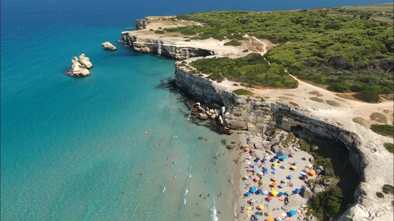 Blick von oben auf türkisblaues Wasser, eine helle Felsküste und eine Badebucht